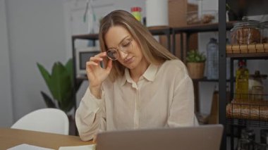 Ofiste gülümseyen bir kadın, gözlük çıkarırken, sarı saçlar, iş kıyafetleri, kapalı mekan dekorları, rafları, bitkileri ve dizüstü bilgisayarı ahşap masa üstünde olan modern iş yerleri..