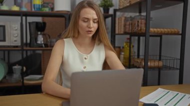 Ofiste çalışan kadın, elleri yüzü dizüstü bilgisayarda endişeli görünüyor. Stres ya da endişeye işaret ediyor. Raflar, belgeler ve bilgisayar gibi işyeri unsurlarıyla çevrili..