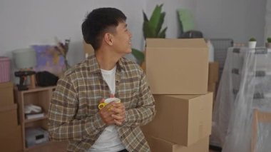 Elinde kahve fincanı tutan genç adam rahat bir oturma odasında kutuları taşırken gülümsüyor ve yeni bir evin heyecanını gösteriyor..