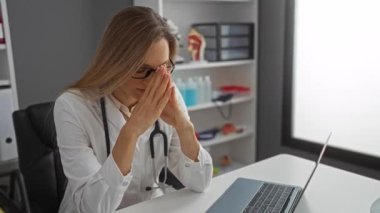 Klinikteki kadın gözlüğü çıkarırken stresli görünüyor, steteskoplu beyaz önlük giyiyor, dizüstü bilgisayarla masada oturuyor, modern iş ortamını gösteriyor..