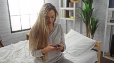 Yatak odasında oturan bir kadın, uzun sarı saçlı, gri pijamalı, kapalı bir yerde oturan, arka planda bitki ve kitaplar olan, pencereden içeri doğru doğal ışık saçan bir kadın..