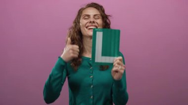 Gülümseyen ve yeşil öğrenci tabelasını tutan bir kadın pembe arka plan duvarına başparmak işareti yaparken