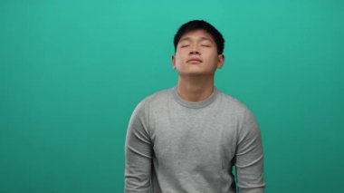 Gözleri kapalı genç adam yeşil arka planda gri süveter giyiyor sükuneti ve memnuniyeti minimalist bir ortamda ifade ediyor..