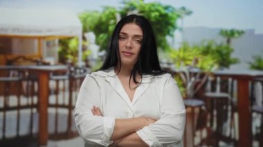 Güneşli bir terasta rahat poz veren siyah saçlı genç bir kadın, bir kahve restoranında yeşilliklerle çevrili, kendine güveni ve sakinliğiyle.