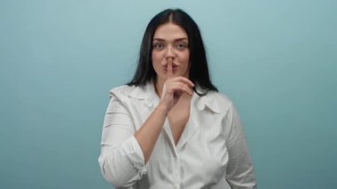 İzole edilmiş mavi arka planda dudakları üzerinde sessizce el kol hareketi yapan kadın, beyaz gömlekli kendine güvenen, sessiz jestler yapan, kıvrımlı bir kadın..