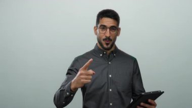 Gözlüklü genç adam, beyaz arka plana karşı tablet tutarken enerjik bir şekilde el kol hareketi yapıyor..