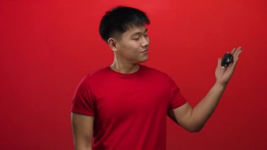 Kırmızı tişörtlü genç adam arabanın anahtarlarını tutuyor. Arka planda izole edilmiş kırmızı bir duvar var..