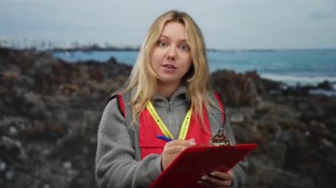 Sahilde arka planda deniz olan, kırmızı yelek giyen ve elinde bir pano tutan gönüllü bir kadın..