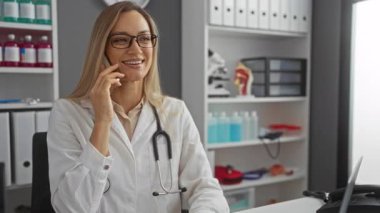 Klinikte telefonla konuşurken gülümseyen bir kadın, gözlük takıyor ve boynunda steteskop olan beyaz bir ceket, etrafında ofis malzemeleri ve tıbbi modeller var..