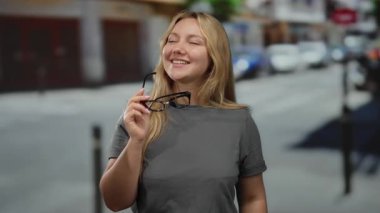 Şehirde gülümseyen bir kadın, elinde gözlükler, sarı saçlar, günlük kıyafetler, açık havada rahat bir şehir ortamını işaret ediyor..