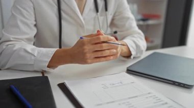 Doktor klinik ofis ortamında tıbbi aletler ve dizüstü bilgisayarla çevrili masada önemli detayların altını çizerek evrak işlerini tartışıyor..