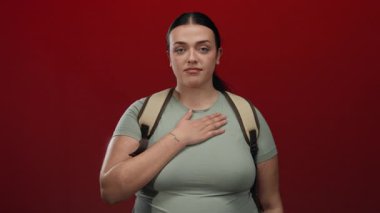 Sırt çantalı genç bir kadın kırmızı duvarın önünde yemin ediyor, eli kalbi ve gözleri kapalı, içtenlik ve bağlılık duygularını iletiyor..