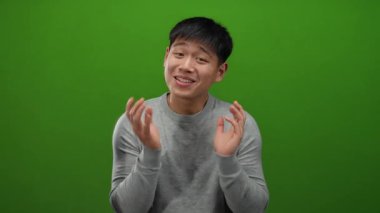 Parlak yeşil arkaplan önünde güven içinde el kol hareketi yapan, günlük gri süveter giyen, duygularını basit bir stüdyo arkaplanına karşı elleriyle ifade eden genç adam.