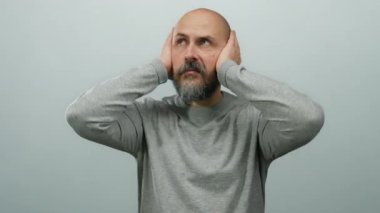 Kulakları elleriyle kapatan, beyaz arka planda hayal kırıklığını ifade eden, günlük gri süveter giyen bir adam..