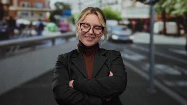 Sarışın ve gözlüklü kendine güvenen genç bir kadın kalabalık bir şehir terasında duruyor, kolları çapraz, açık bir şehir ortamında siyah bir ceket giyiyor..