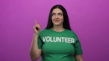 Yeşil gönüllü tişörtü giyen bir kadın canlı pembe bir arkaplanı işaret ederek yukarı doğru gülümsüyor. Renkli, izole bir ortamda pozitiflik ve aktivizm sergiliyor..