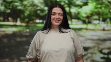 Genç bir kadın güvenli bir şekilde açık hava parkında gülümsüyor yemyeşil ve güneş ışığıyla serin bir atmosfer yaratarak neşesini ve güvenini gösteriyor..