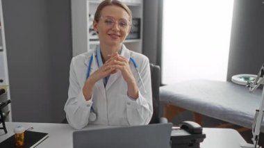 Klinikteki kadın doktor, kollarını kavuşturarak gülümsüyor, beyaz önlük üzerine steteskop takıyor, tıbbi malzemeler ve parlak pencere altyapısı ile çevrili..