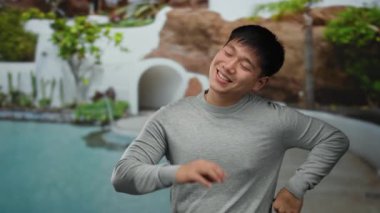 Genç bir adam açık havada güneşli gökyüzünün altında güneşli bir otel havuzunda gülüyor. Arkaplanda yemyeşil yemyeşil bir hava yayıyor..
