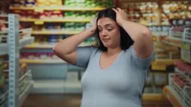 Mavi tişörtlü, uzun siyah saçlı kadın süpermarket koridorunda duruyor. Renkli rafların arasında elleri başının üstünde duruyor..