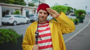 Sarı yağmurluklu ve kırmızı bereli genç adam şehir sokaklarında oltayla selamlıyor. Açık hava cazibesini somutlaştırıyor..