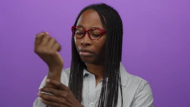 Bileği ağrıyan, gözleri kapalı, mor bir arka plana bakan, gözlüklü ve çizgili bir gömlek giyen bir kadın izole edilmiş bir stüdyoda rahatsız edici bir görüntü sergiliyor..