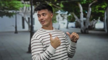 Genç adam arka planda ağaçlar ve binalar olan bir caddede başparmak kaldırıyor, çizgili bir gömlek giyiyor ve kendine güvenerek gülümsüyor, pozitif tavır sergiliyor..