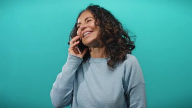 Gülümseyen, telefonda mutlu bir şekilde konuşan, kıvırcık saçlı, canlı mavi bir duvara bakan, izole bir ortamda neşe ve bağlantı gösteren bir kadın..