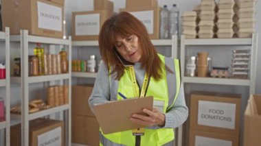 Bağış merkezinde çalışan, panoya yazı yazan, yemek, kutu ve raflarla çevrili telefonla konuşan, gönüllü yelek giyen son sınıf İspanyol bir kadın..