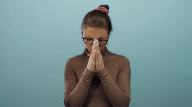 Gözlüklü ve kahverengi bluzlu genç bir kadın, izole edilmiş mavi arka plan duvarına sıkıca sarılmış ellerle dua ediyor.