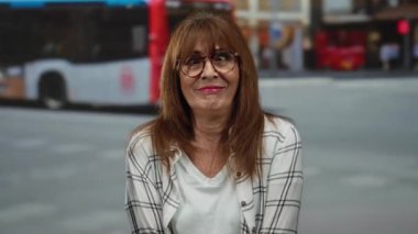 Arka planda bulanık otobüsler ve ekose gömlek ve gözlüklerle işlek bir şehir caddesinde neşeli bir şekilde gülümseyen İspanyol son sınıf öğrencisi kadın..