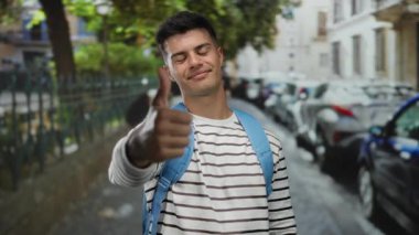 Şehir caddesinde başparmağıyla gülümseyen genç bir adam, çizgili bir gömlek ve mavi bir sırt çantası, park etmiş arabalar ve ağaçlarla çerçevelenmiş, dışarıda pozitiflik yayıyor..