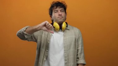 Sarı ses geçirmez kulaklık takan İspanyol bir adam turuncu bir arka plana başparmak göstererek canlı bir şekilde onaylamama hissini yakalıyor..