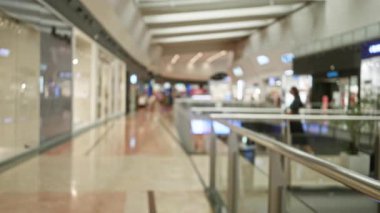 Bulanık alışveriş merkezi koridorunda insanlar yürüyor, ışıkların yönünü değiştiriyor, ve modern perakende mağaza cepheleri işlek bir ticari atmosfer yaratıyor.