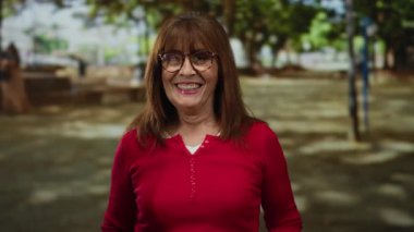 Kıdemli İspanyol kadın, güneşli bir açık hava parkı ortamında gülümseyip dişlerini gösteriyor. Keyif ve sıcaklık saçıyor. Günlük tarzıyla ve doğal güzelliğiyle..