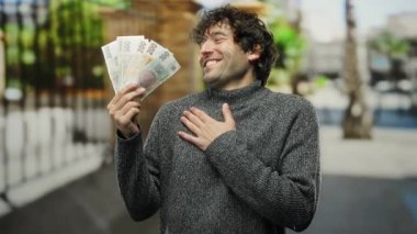 Dışarda gülümseyen İspanyol bir adam göğsünde bir el ile Czech Koruna banknotlarıyla şehrin arka planında mutluluğu ve finansal başarıyı işaret ediyor..