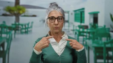 Gözlüklü yaşlı bir kadın, bir restoranın terasında, güneşli bir caddede, yeşil sandalyelerle çevrili bir yerde şaşkınlıkla kendini işaret ediyor..