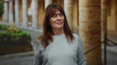 Kıdemli İspanyol kadın, arka planda canlı yeşillik ve antik mimari ile şehir merkezindeki çevre seslerini dikkatle dinliyor ve gülümsüyor..