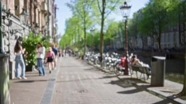 Amsterdam, Hollanda 'da kanalların kenarında yürüyen, kadınları rahat ve manzaralı bir şehir ortamında, yolu çevreleyen yemyeşil ağaçlar ve bisikletlerle yakalayan insanların odaklanmamış görüntüsü..