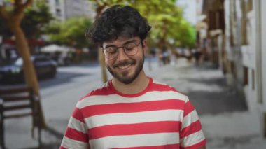 Genç adam, sakallı ve gözlüklü canlı bir sokakta arka planda ağaçlar ve arabalarla gülümsüyor, çizgili bir gömlek giyiyor..