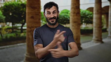 Sakallı bir adam eski Roma 'da mutlu bir şekilde hareket ediyor. Arka planda bulanık sütunlar ve yeşillikler var. Gülümsüyor ve işaret ediyor..