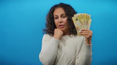 Romen banknotlarını mavi arka planda tutan kadın para ve ifade odaklı mali konsept tasvir ediyor, günlük süveter giyiyor, izole edilmiş ve gözlemliyor.