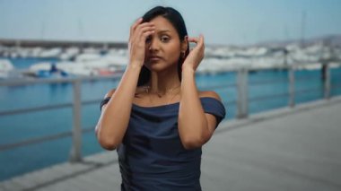 Deniz kenarında sakin bir ifadeyle duran kadın, deniz esintisini hissediyor, mavi bir elbise giyiyor, marinada gün boyunca arka planda tekneleri olan bir kadın..