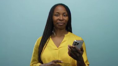 Mavi arka planda duran akıllı telefondan gülümseyen, sarı gömlekli, kameraya doğru bakan bir kadın güven pozitifliğini ve bağlantıyı temsil ediyor..