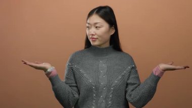 Avuçlarını kahverengi bir duvara dayamış, gri bir kazak giyen, seçimini ya da sunumunu sakin bir ifadeyle yapan bir kadın..