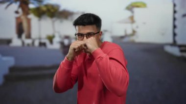 Kırmızı tişörtlü genç adam güneşli bir şehirde beyaz binalar ve palmiye ağaçlarıyla çevrili bir sokakta yumruklarıyla şakacı pozlar veriyor..