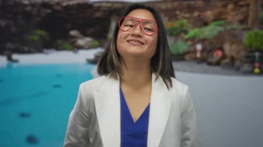 Açık hava tatil köyünde turkuaz havuzunda parlak gözlüklü beyaz ceketli bir kadın gülümsüyor..