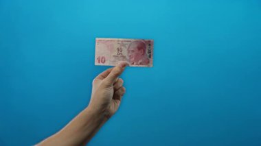 Türk lira banknotunu tutan adam mavi arka planda izole edilmiş, minimalist bir ortamda para birimi ayrıntılarını gösteriyor..