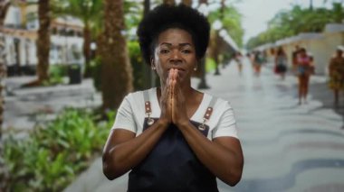 Yoğun şehir caddesinde açık havada önlük giyip dua eden bir kadın, kentin yeşillik ve arka plandaki insanlarla çalkantılı ortamına dinginlik ve maneviyat katıyor.