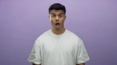 Genç adam, canlı bir mor arkaplan üzerine izole edilmiş, sıradan beyaz bir gömlek giyen, yüzün yanında elleri olan bir dizi duygu sergileyen, şaşırdığını ifade ediyor..